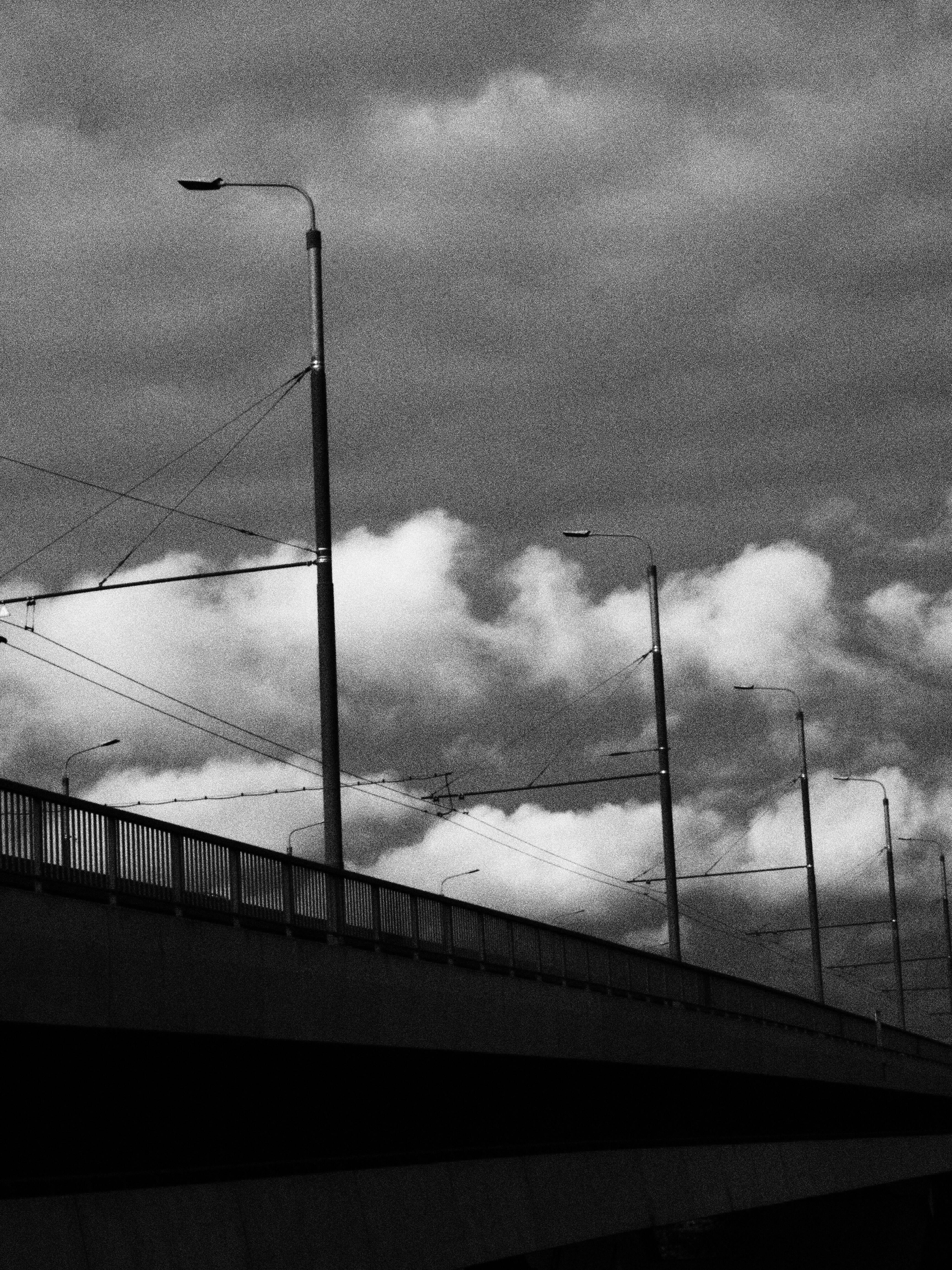 Dramatic black and white photo of a bridge with lampposts under cloudy skies, capturing urban melancholy.