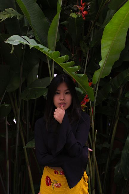 A thoughtful young woman stands elegantly surrounded by lush tropical leaves.