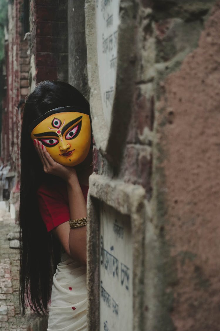 Teenager Girl In Yellow Durga Mask Peeking Behind A Wall On A Street