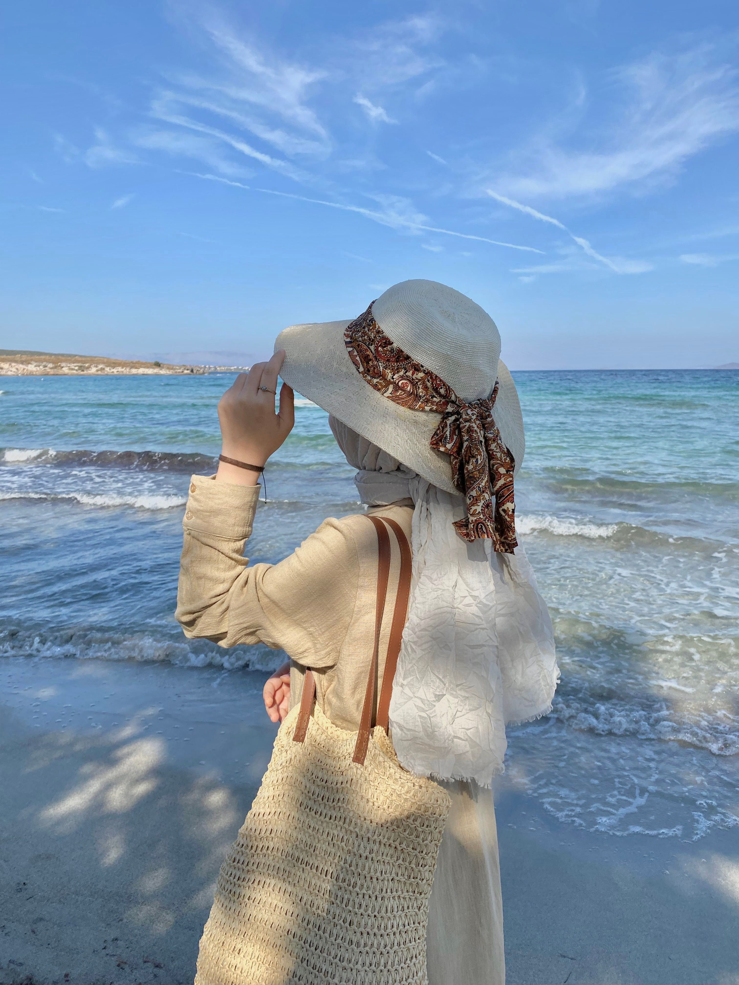 A fashionable woman wearing a hat and shawl stands by the sea, enjoying a sunny day at the beach.
