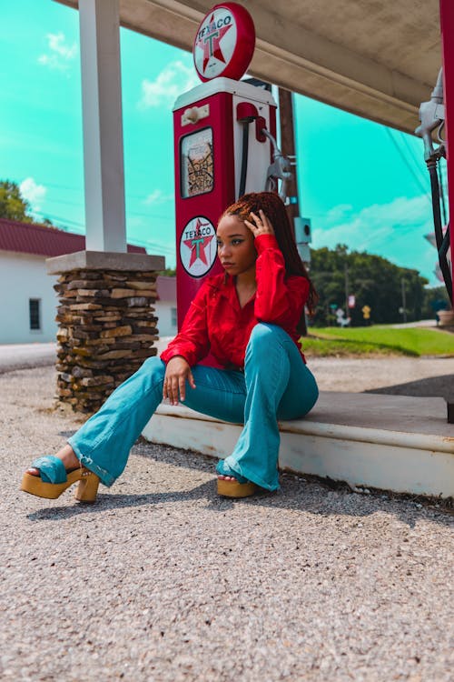 Young Woman in a Red Blouse and Jeans Sitting at a Gas Station 