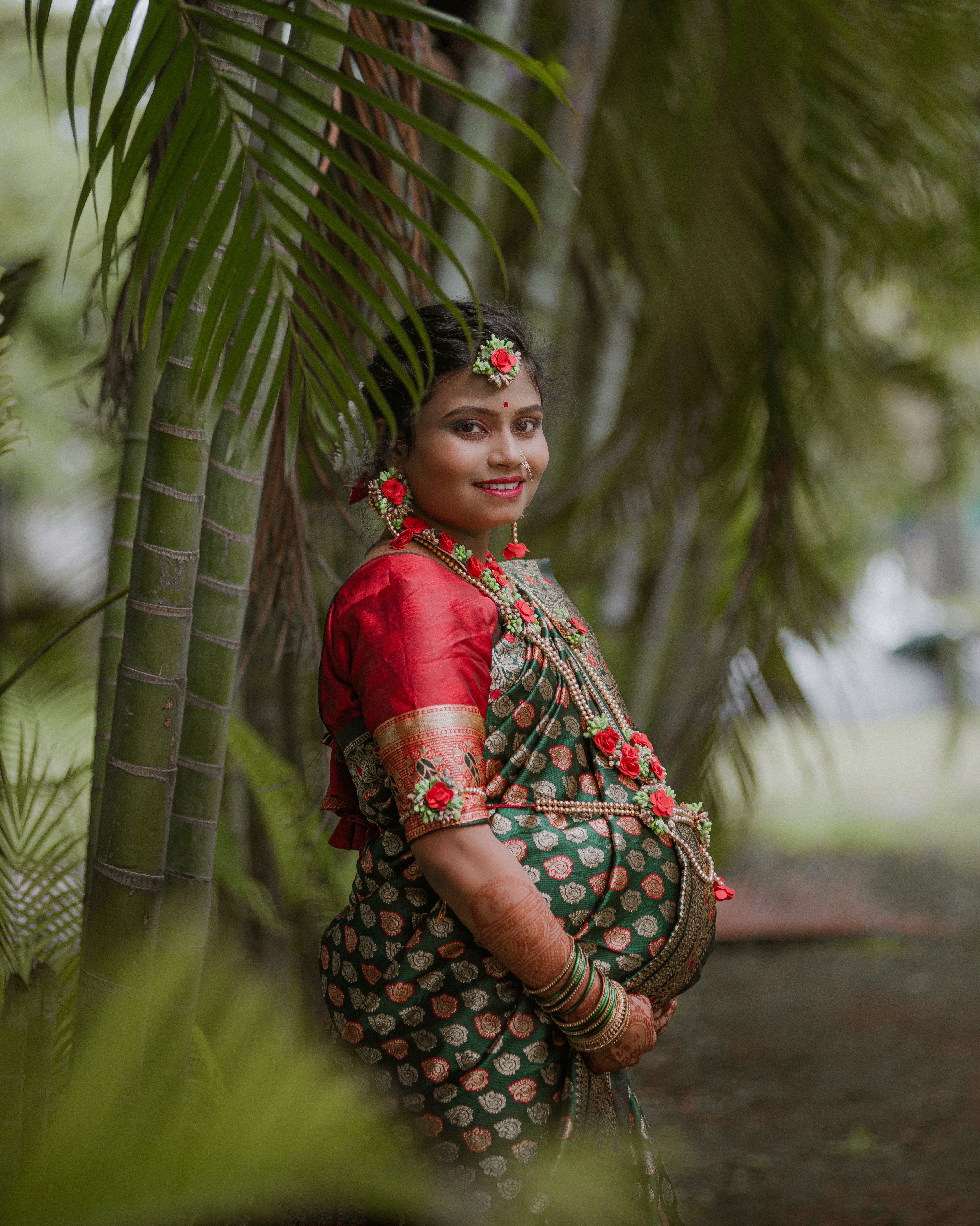 Smiling pregnant woman in traditional Indian clothing standing outdoors surrounded by lush greenery.