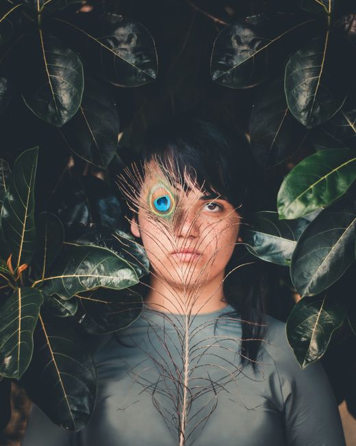 A woman poses with a peacock feather by her face surrounded by large dark leaves.