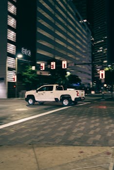 A white pickup truck drives through a dimly lit urban street at night, isolated with blurred motion.