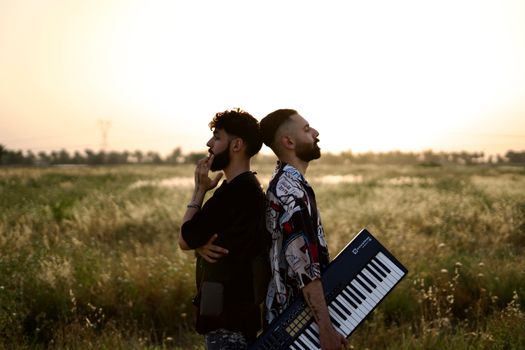 Two men standing back to back in a field, one holding a keyboard, during sunset.