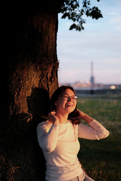 Free A woman with glasses smiles while sitting by a tree during sunset in a park. Stock Photo