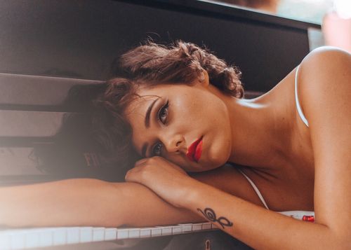Free A thoughtful woman in a relaxed pose with striking red lipstick rests her head on a piano. Stock Photo