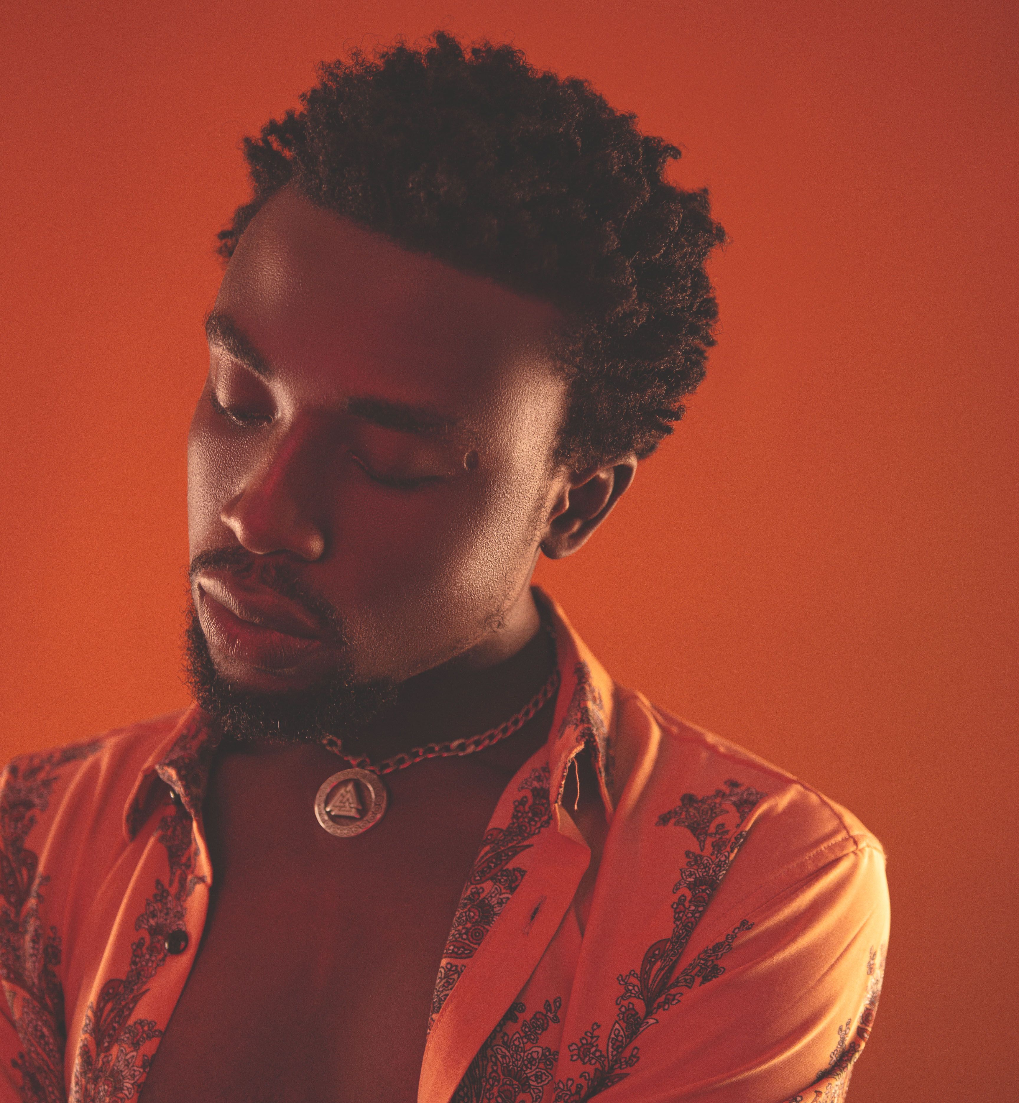 Close-up portrait of a man with a beard and necklace against an orange background. Serious expression, vibrant color.