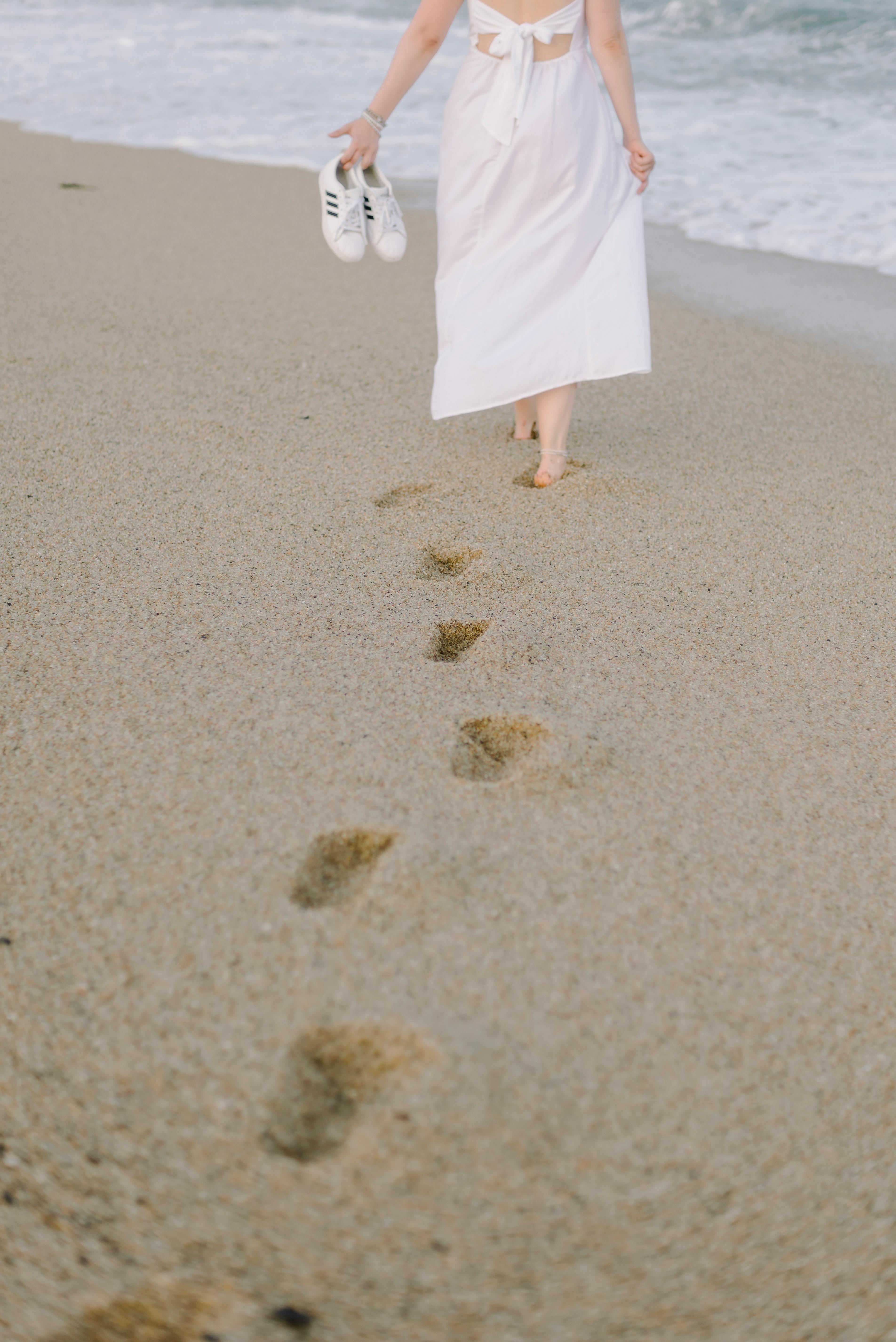 A serene scene of a woman walking barefoot on a sandy beach in a white dress carrying sneakers.