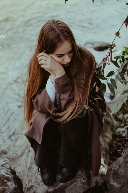 Young woman sits by a river, deep in thought, amidst nature's solitude.
