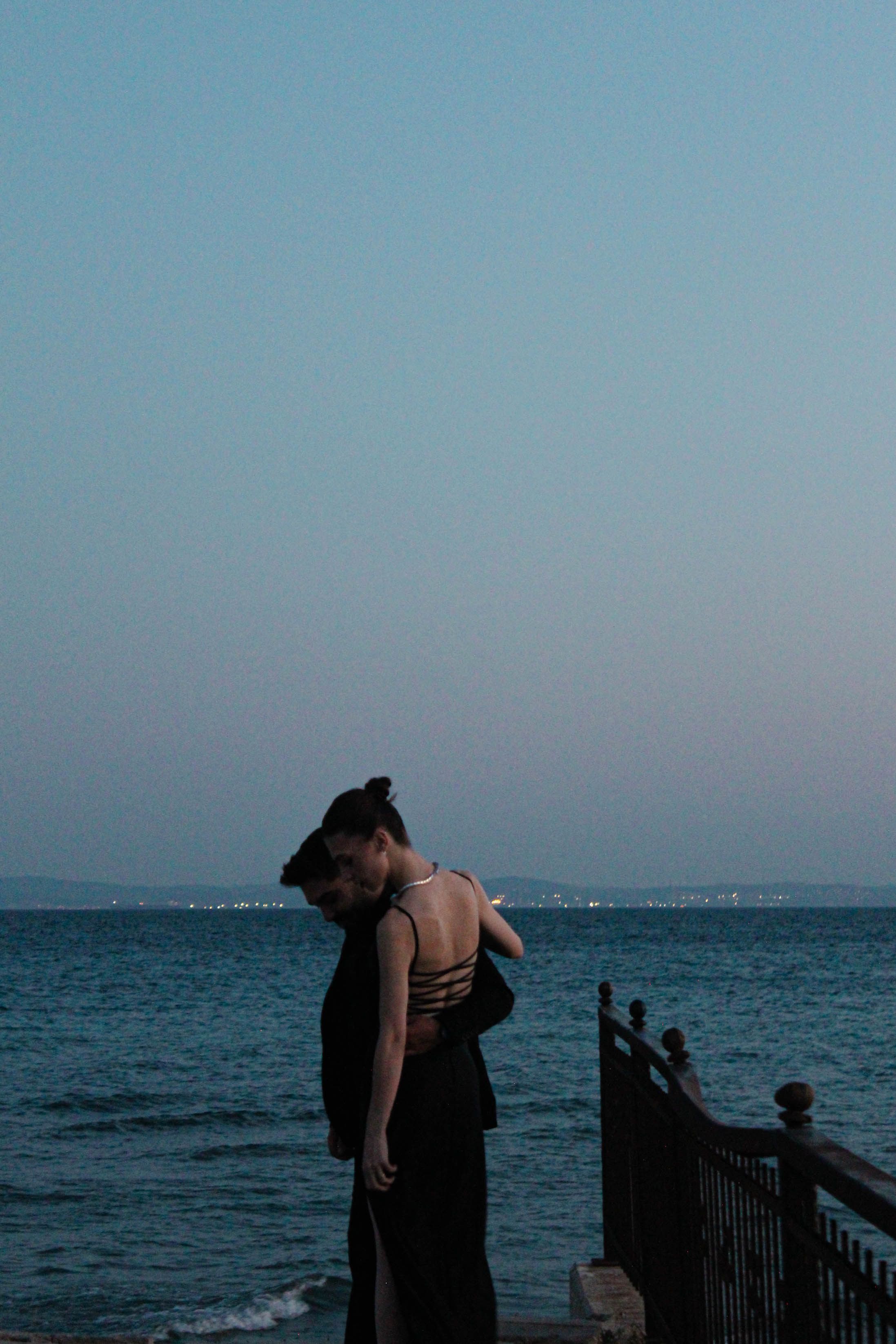 A couple in stylish black attire embraces by the sea during twilight, exuding romance.