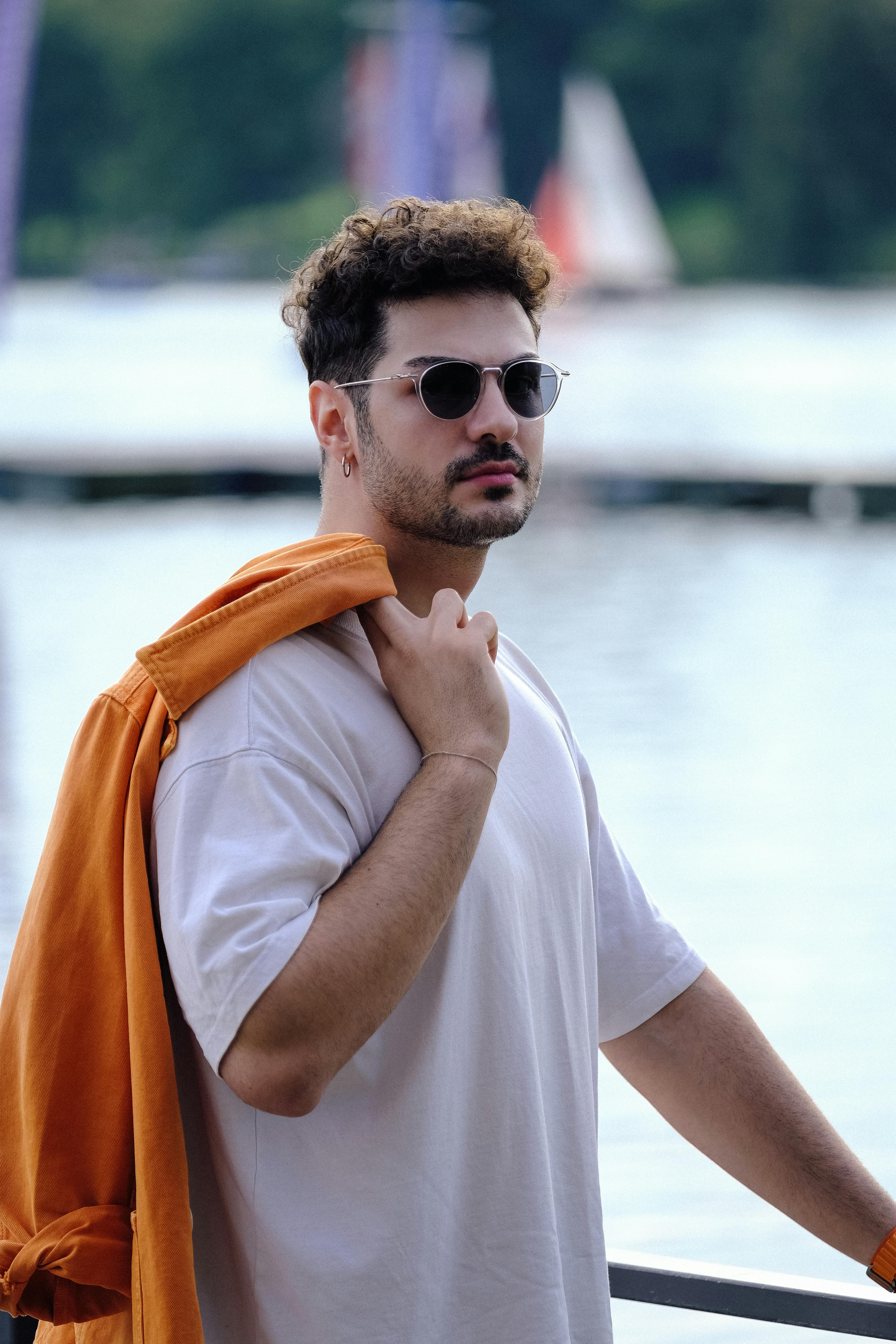 Fashionable man wearing sunglasses and an orange jacket posing by the lake in summer.