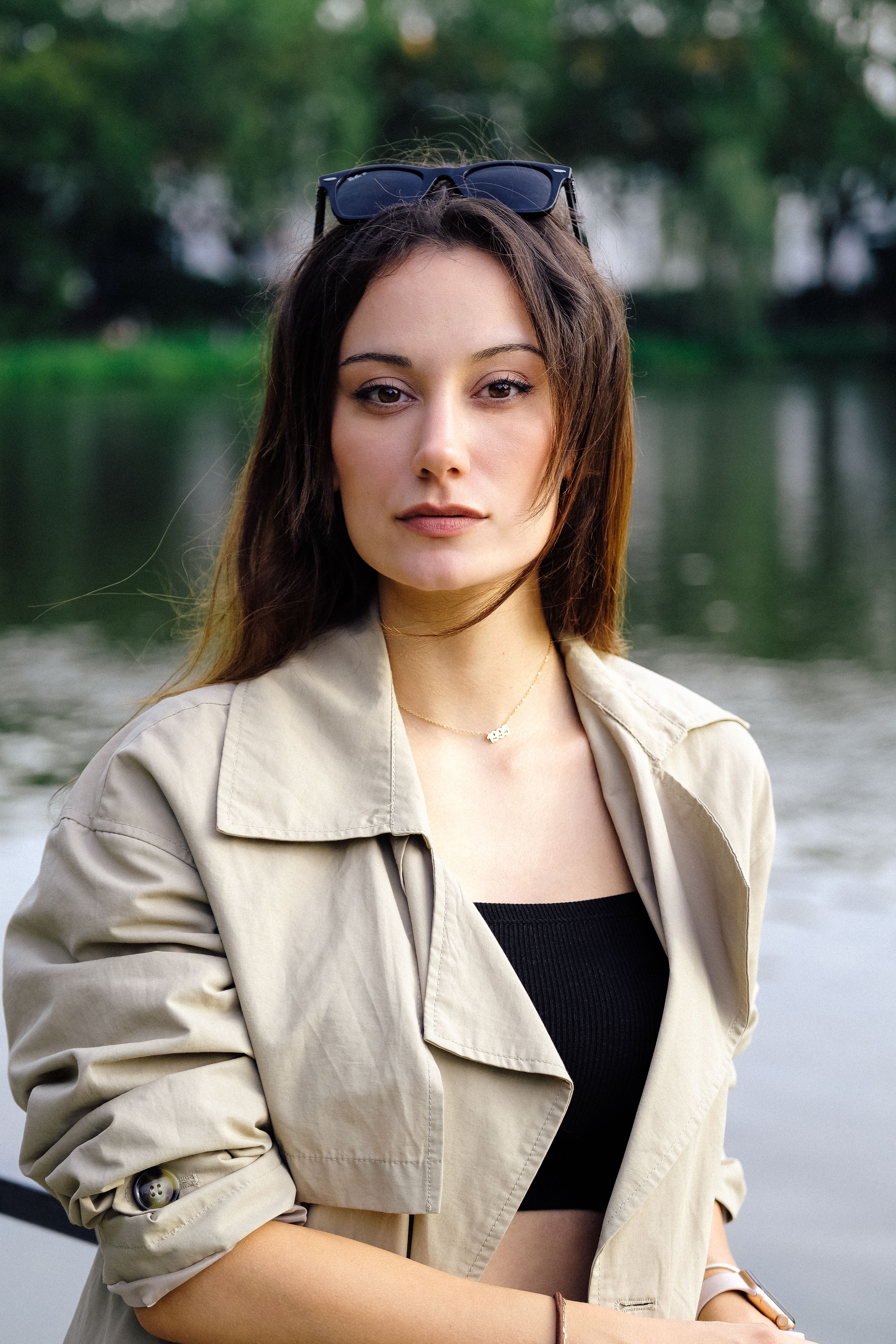 Portrait of a young woman with long hair and sunglasses in a trench coat by the water.