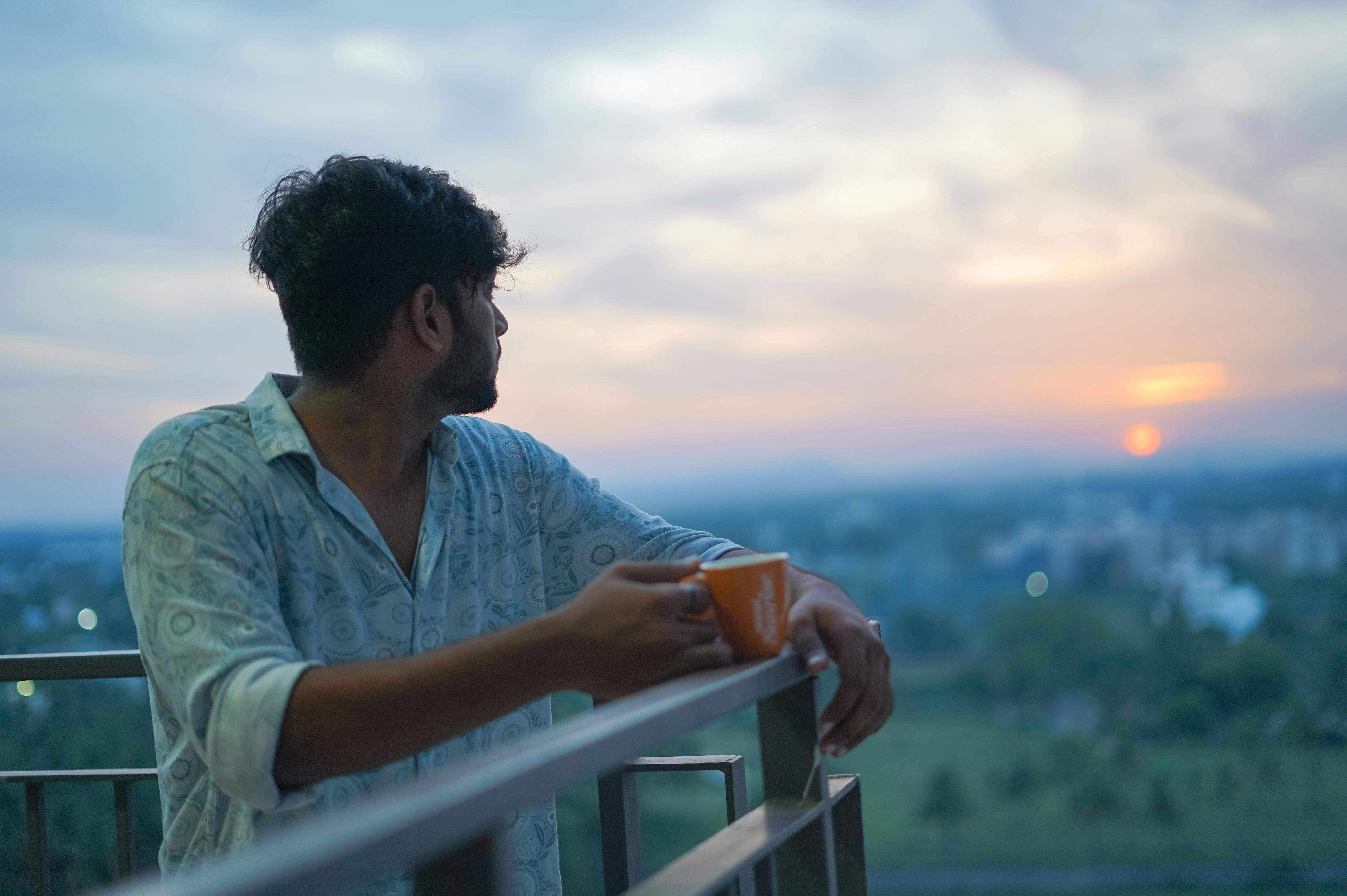 A young man enjoying a peaceful sunrise while holding a cup of coffee on a balcony.