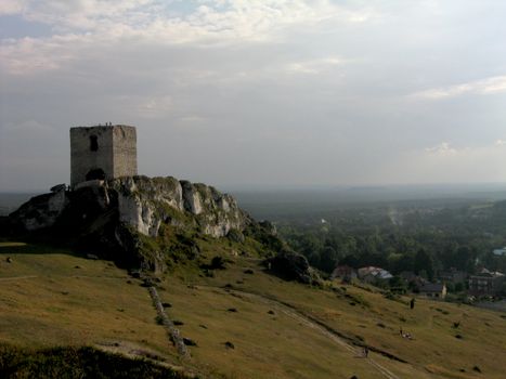 A historic tower stands atop a rocky hill with scenic rural Polish landscape in the background.