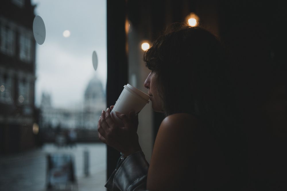 Femeie bând cafea lângă o fereastră, cu o stradă europeană neclară în fundal