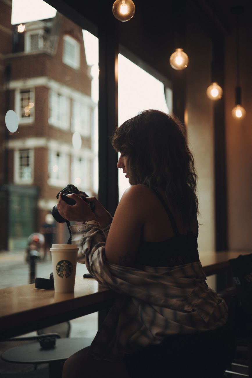 A woman sitting by the window in a café, holding a camera and enjoying her coffee.