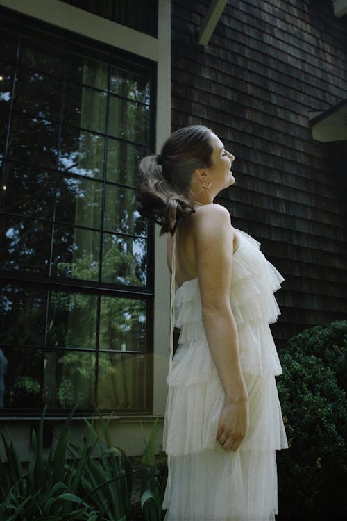 Smiling Woman in White Dress Standing by House Window