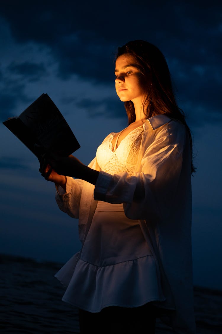 A Woman Standing In The Water With A Book Illuminating Her Face