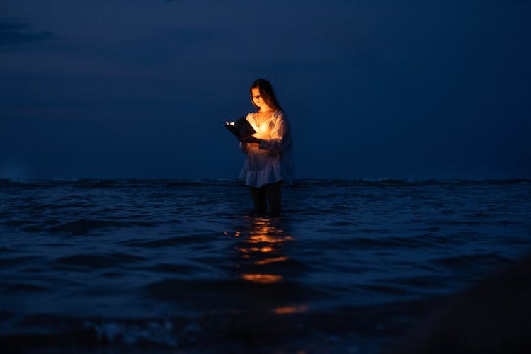 A Woman Standing In The Water With A Book Illuminating Her Face