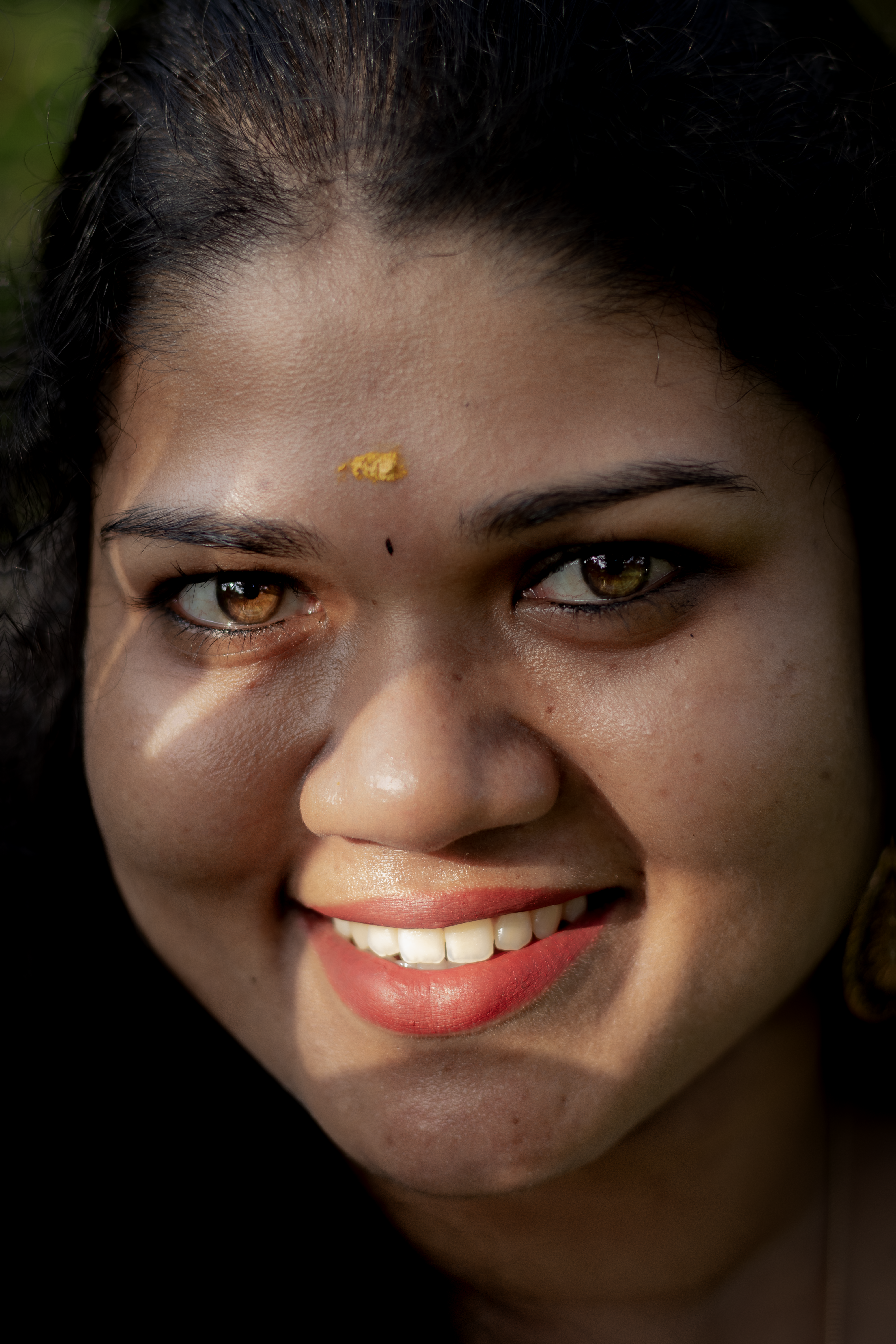 A close-up portrait of a smiling woman with black hair and traditional bindi, captured outdoors.