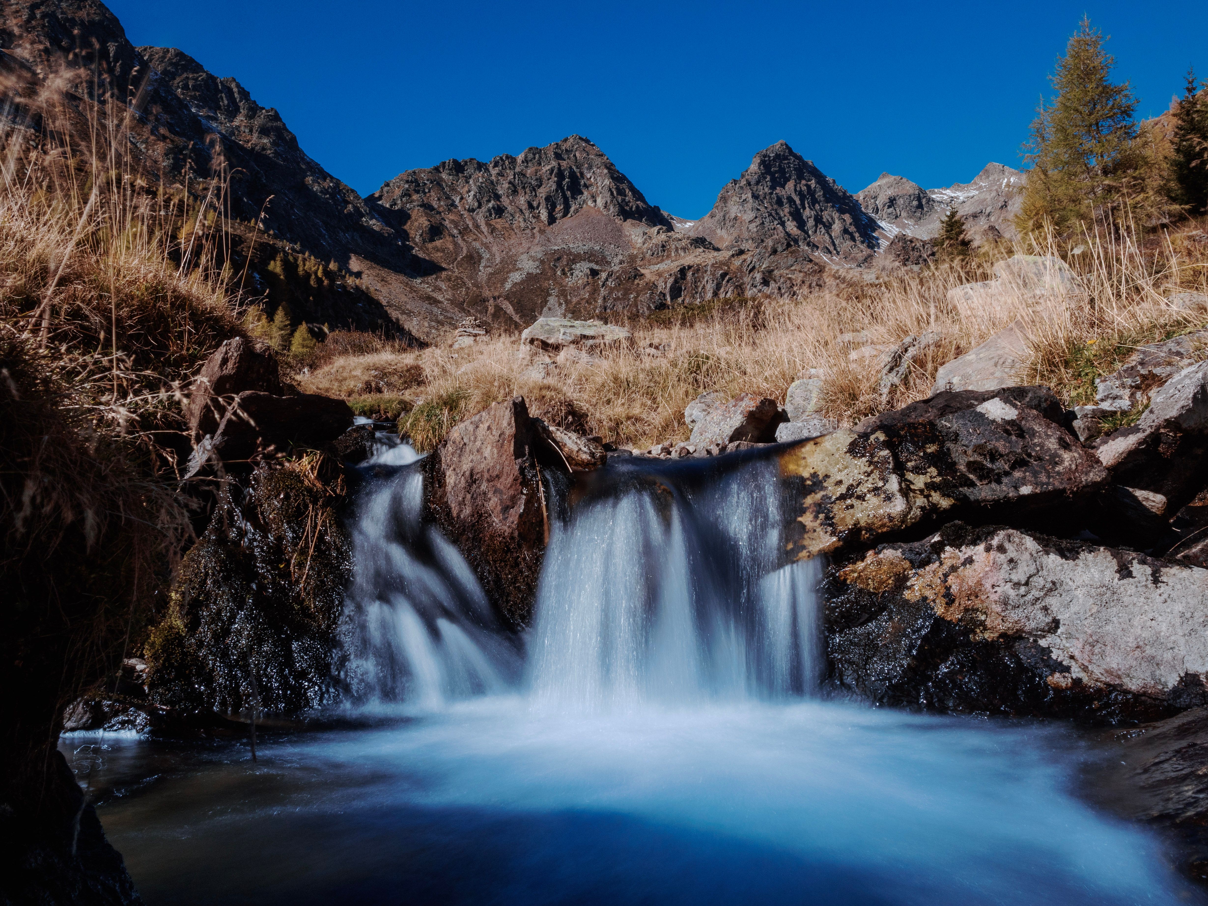 A tranquil waterfall cascading down rocks with towering mountain peaks under a clear blue sky.