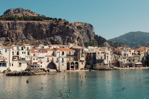 Free Swimmers enjoy the water with Cefalù's historic seaside buildings and rocky cliffs in Sicily, Italy. Stock Photo