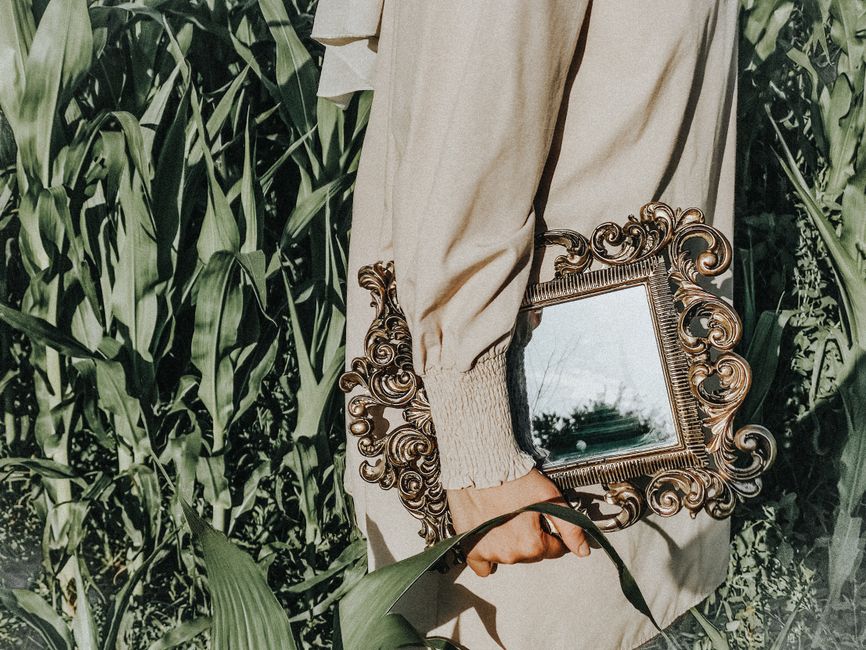 A woman in vintage attire carries an ornate mirror in a lush cornfield, embodying a summer vibe.