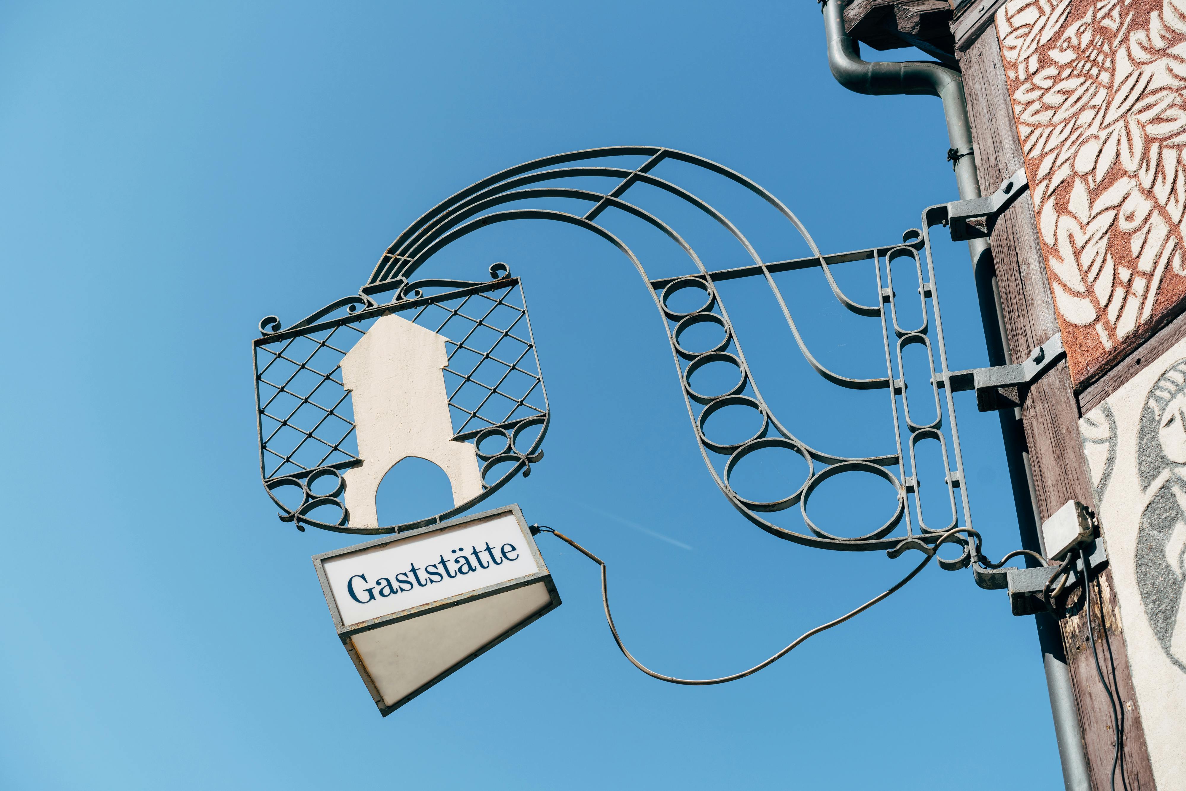 Artistic metal restaurant sign reading 'Gaststätte' against clear blue sky.