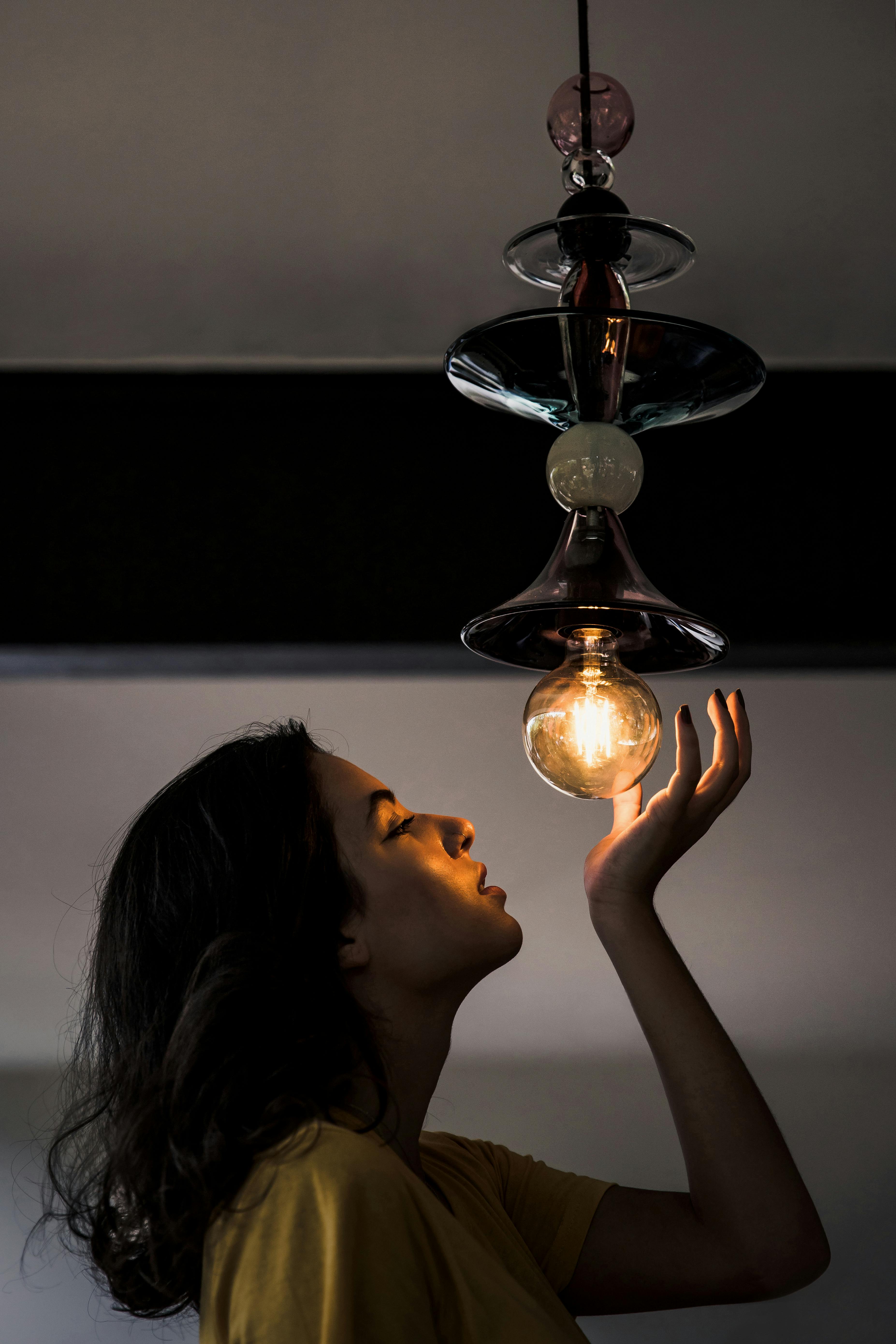 一个女人凝视着设计独特的灯具中发光的灯泡，神情专注，营造出喜怒无常的氛围