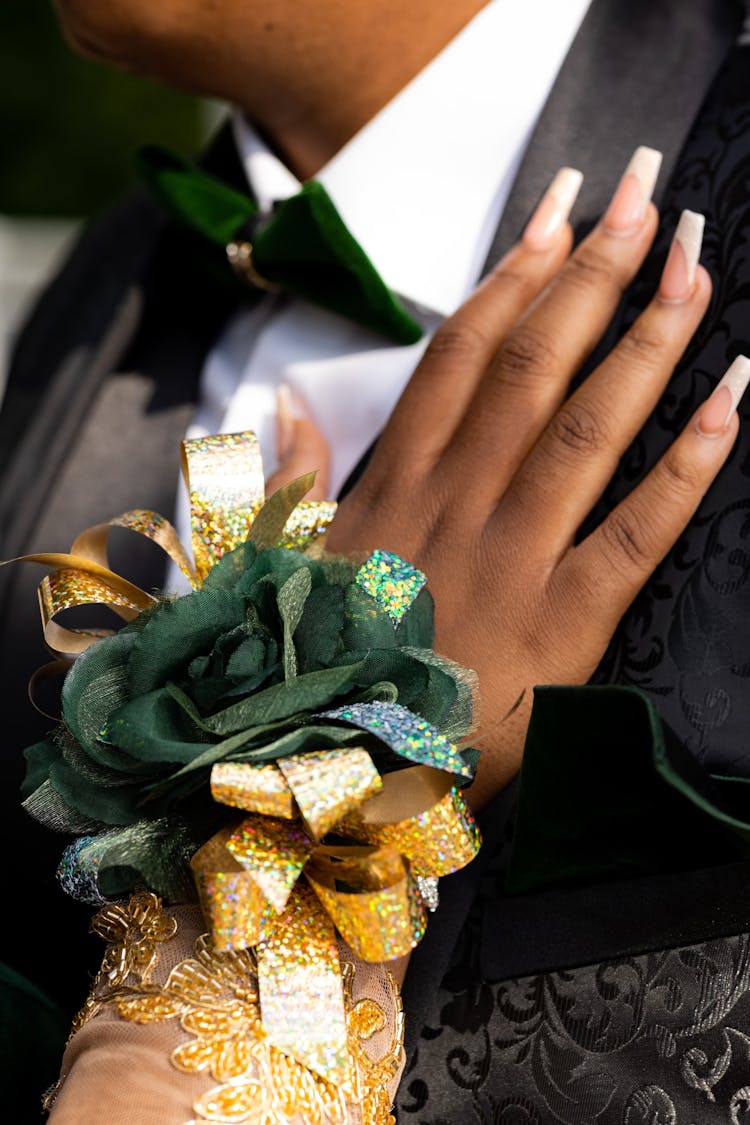 Hand With Hybrid Nails Wearing Gold Bow With Green Flower Touching Person In Suit