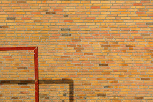 A red metal frame casting a shadow against a textured yellow brick wall with a repeating pattern.