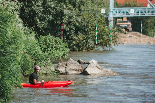 Free A man in a red kayak paddles through a river with lush greenery and rocks on a sunny day. Stock Photo