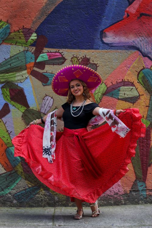 Free Woman in traditional Mexican dress and sombrero dancing against a colorful mural backdrop. Stock Photo