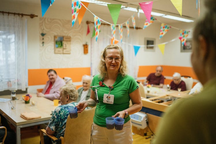 Caregiver Serving Food For Elderly People In Retirement House