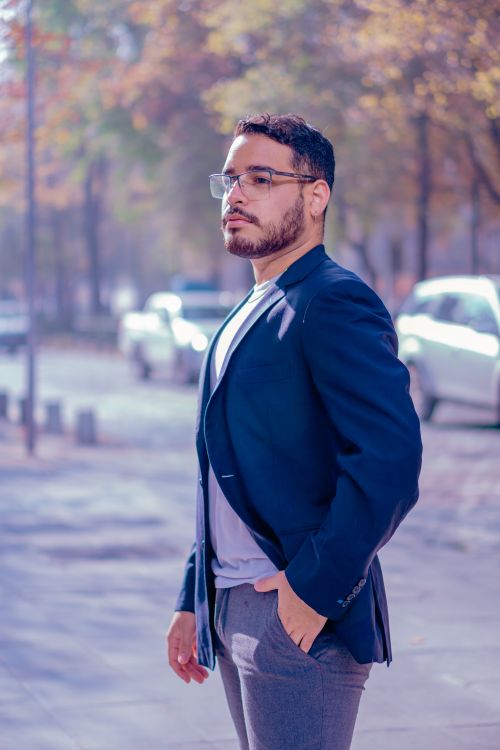 Free A confident man in smart attire standing on a city sidewalk surrounded by autumn foliage. Stock Photo