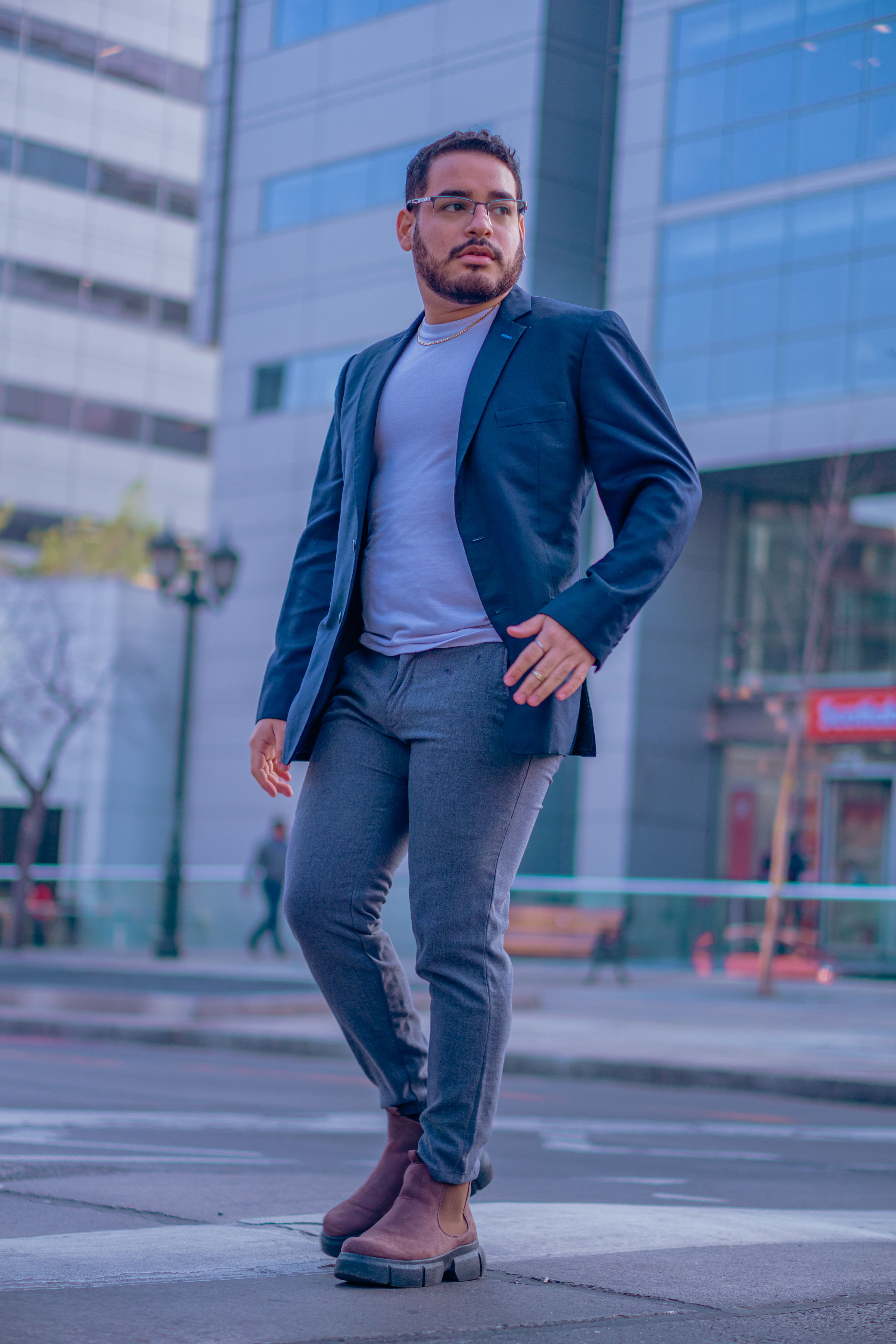 A fashionable man with a beard and glasses walks confidently in a cityscape, showcasing modern urban fashion.