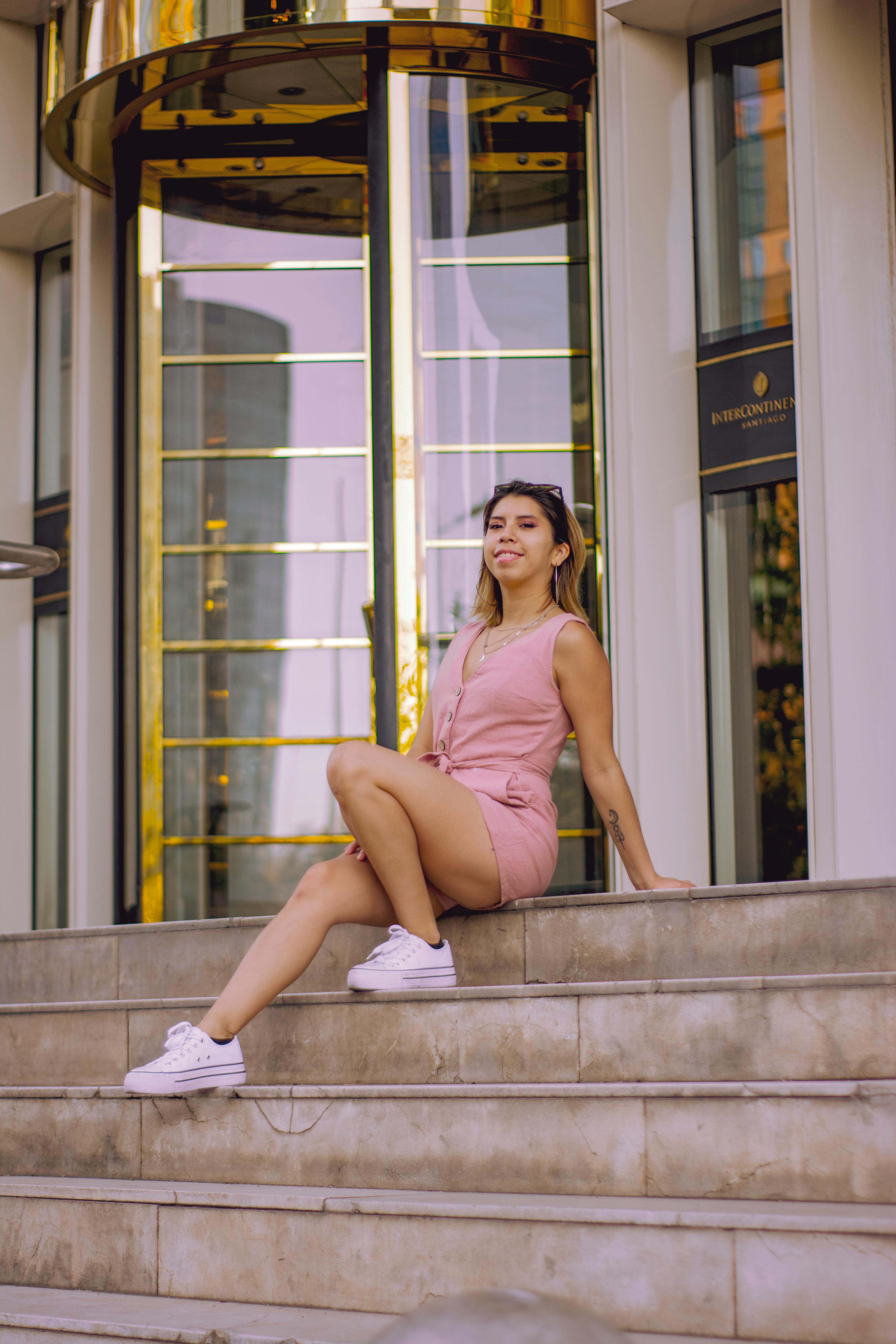 A stylish woman poses in a pink dress on the steps outside a hotel, showcasing urban fashion.