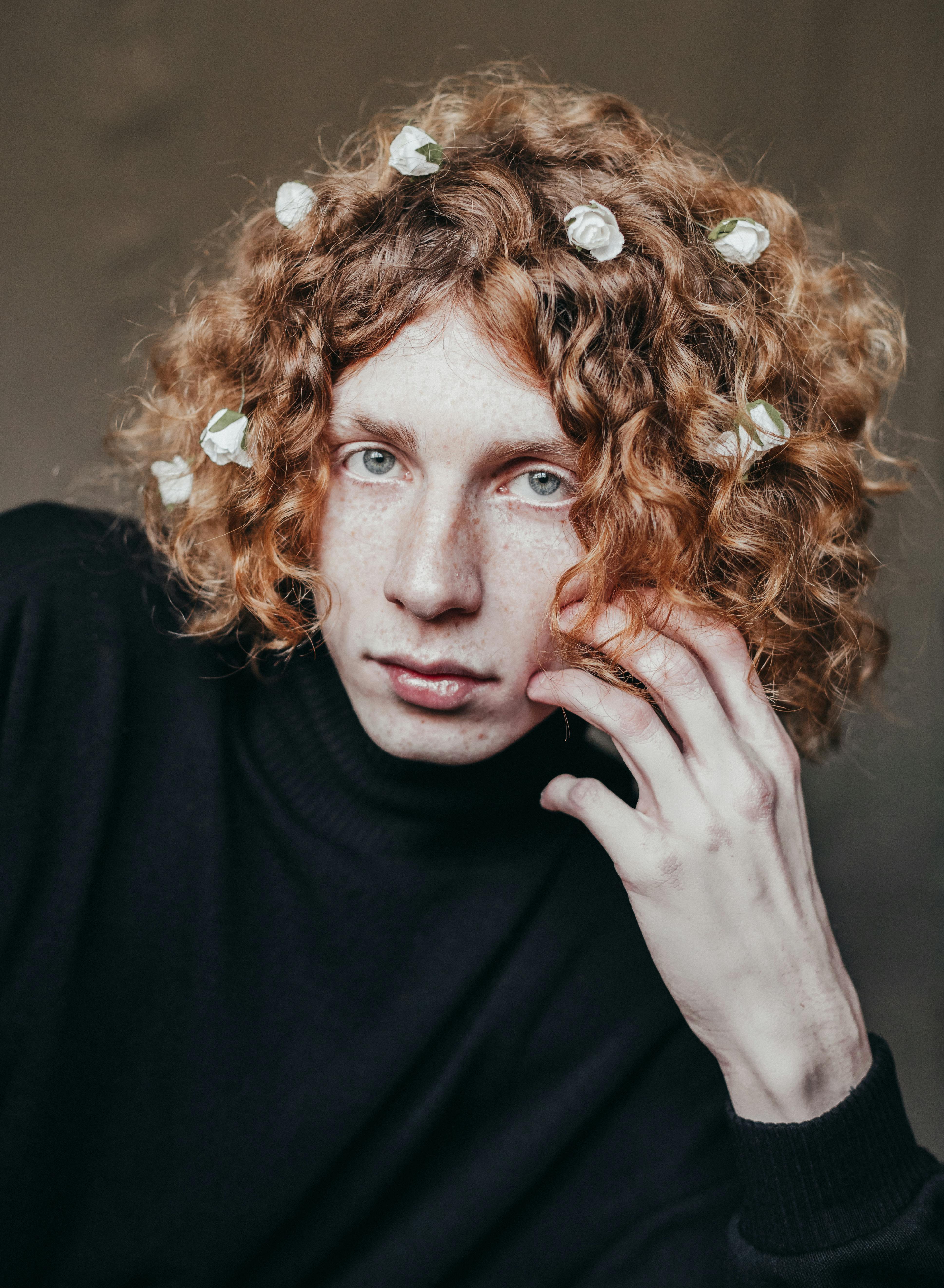 Artistic portrait of a young man with curly hair adorned with flowers, wearing a black turtleneck.
