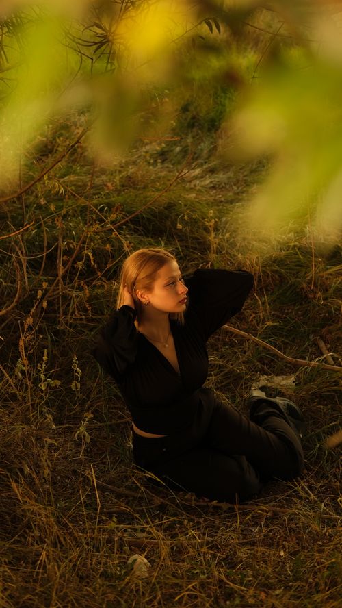 Free A young woman in a black outfit poses thoughtfully in a natural outdoor setting, illuminated by warm sunlight. Stock Photo