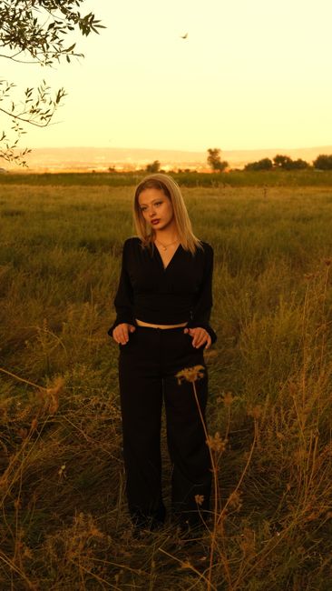 An elegant woman poses in a meadow at sunset, wearing a black crop top and pants.