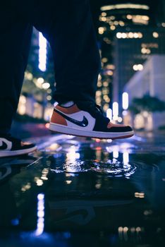 Close-up of sneakers reflecting in a puddle on an urban city street at night.