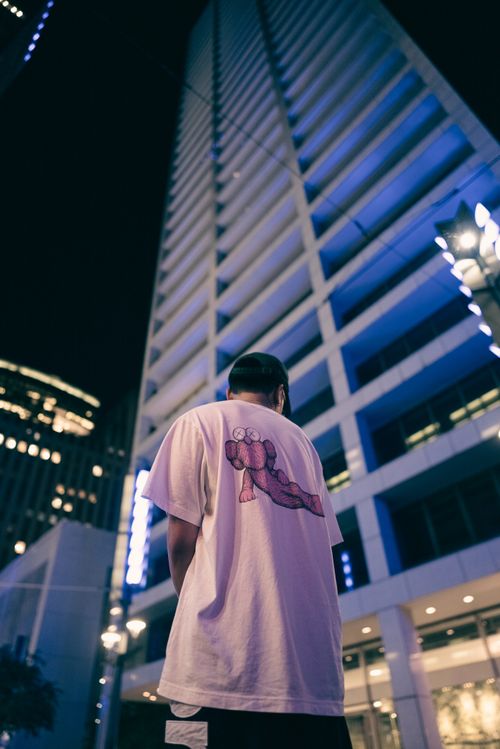 Free Man in white t-shirt stands beneath illuminated skyscraper at night, capturing urban essence. Stock Photo