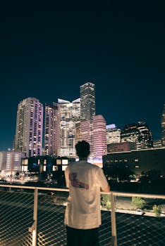 Man in white t-shirt observes illuminated skyscrapers at night from a railing