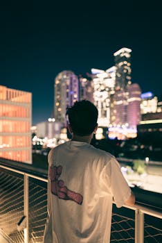 Back view of a man standing on a terrace, overlooking illuminated skyscrapers at night.
