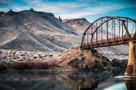 一座金属桥横跨在平静的河流上，背景是雄伟的山脉，景色迷人