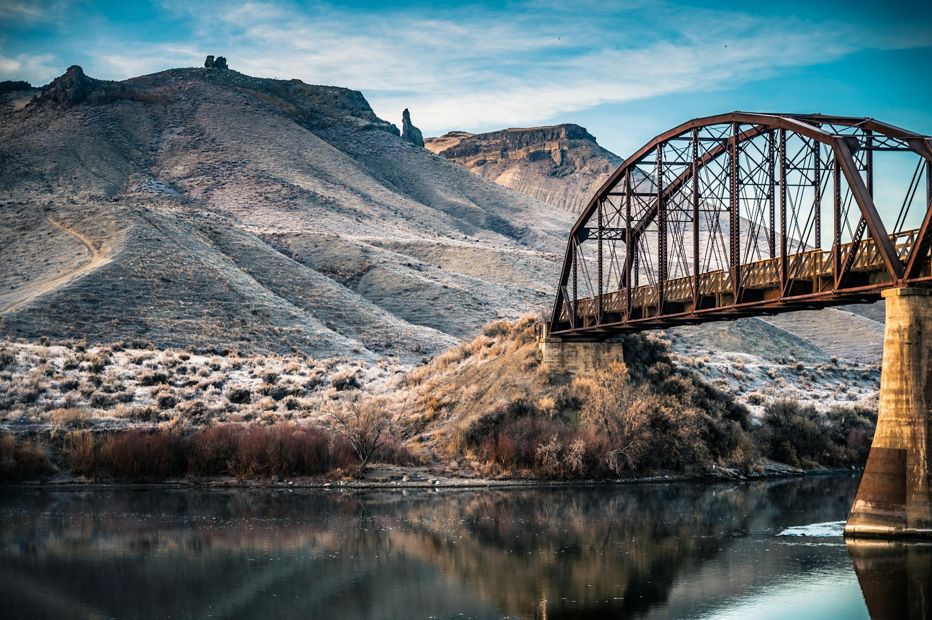 Captivating landscape of a metal bridge spanning over a calm river with majestic mountains in the backdrop.