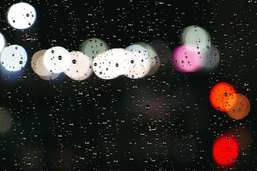 Raindrops on window glass with colorful bokeh lights in the background, creating a dreamy night scene.