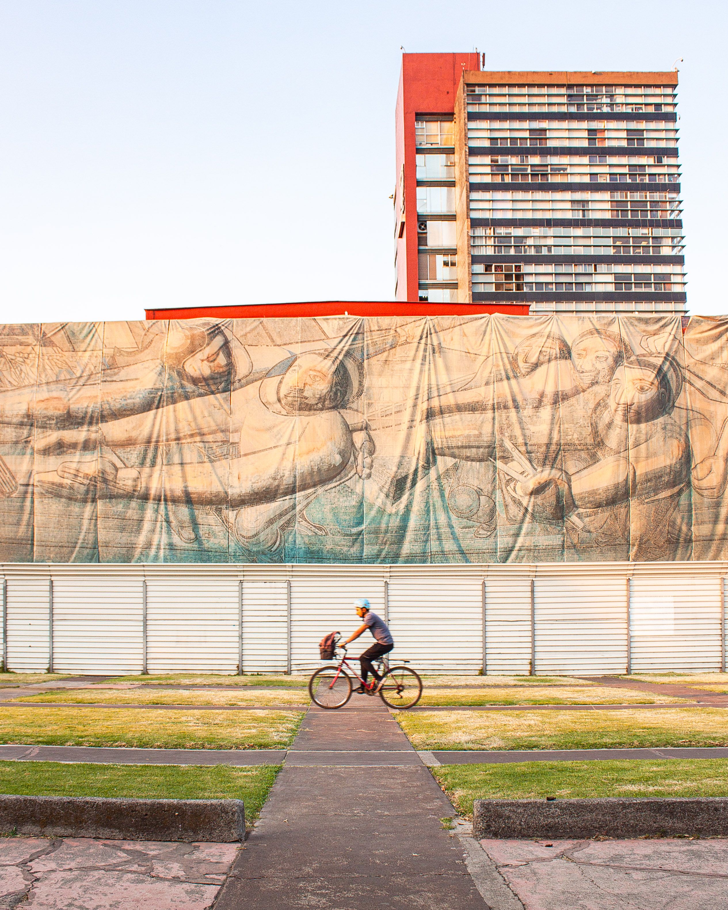 A cyclist rides past a historic mural with a modern high-rise in Mexico City, showcasing urban art.