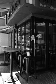 A black and white photo of an Italian pizzeria with stools and tables outside, reflecting a classic ambiance.